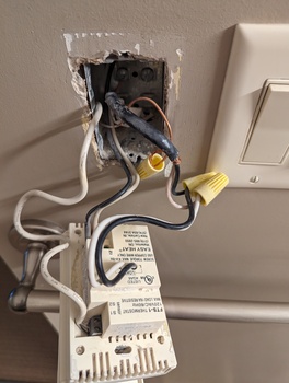 A beige thermostat pulled out of the wall; black and white wires run into it.  At the top of the device, bottom of the photo, a black and white small gauge wire run in from the thermistor as a temperature sensor.  In the middle, heavier wires carry the AC load into the resistive heating of the floor.  And at the bottom, black and white wires provide the AC power input.