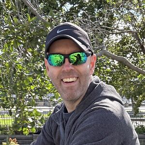 Male smiling face with short trimmed full beard, wearing a black running cap and green reflective sunglasses