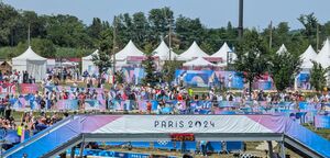 Outdoor photograph of steel barricades and a pedestrian bridge.  The barricades are decorated with pink and blue "Paris 2024" signage, and people are navigating the barricades in thee background.